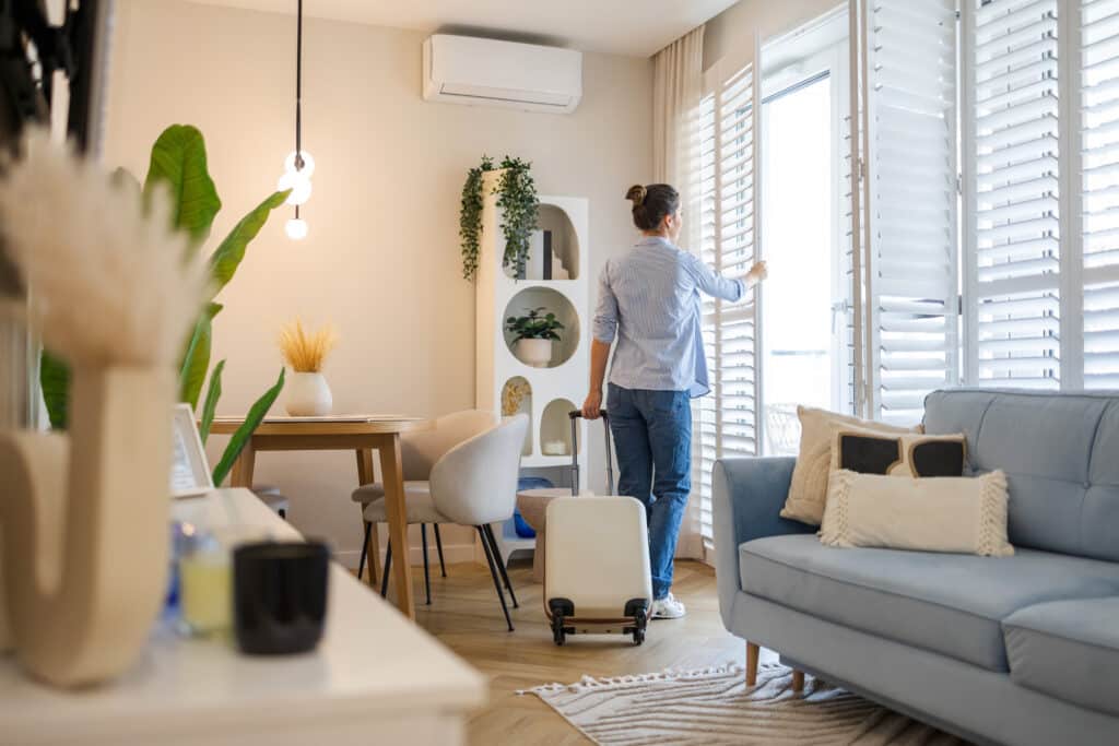 Femme avec valise ouvrant des volets blancs dans un appartement lumineux. Salon moderne, canapé bleu, table et plantes.
