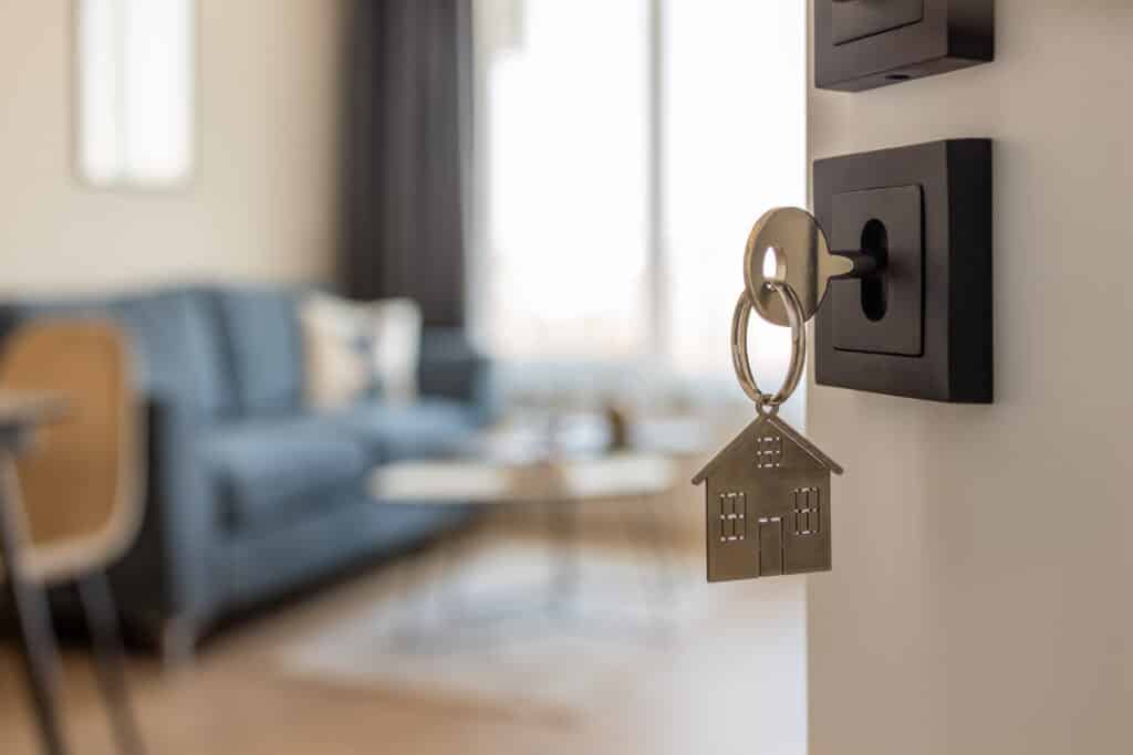 Clé de maison : votre accès à la propriété et au confort. Clé et porte-clés maison dans une serrure noire moderne, avec salon lumineux et canapé bleu flou en arrière-plan.