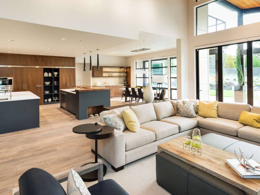 Salon et cuisine ouverts, design moderne. Canapé beige en L, îlots de cuisine gris foncé, meubles en bois. Grandes baies vitrées lumineuses.