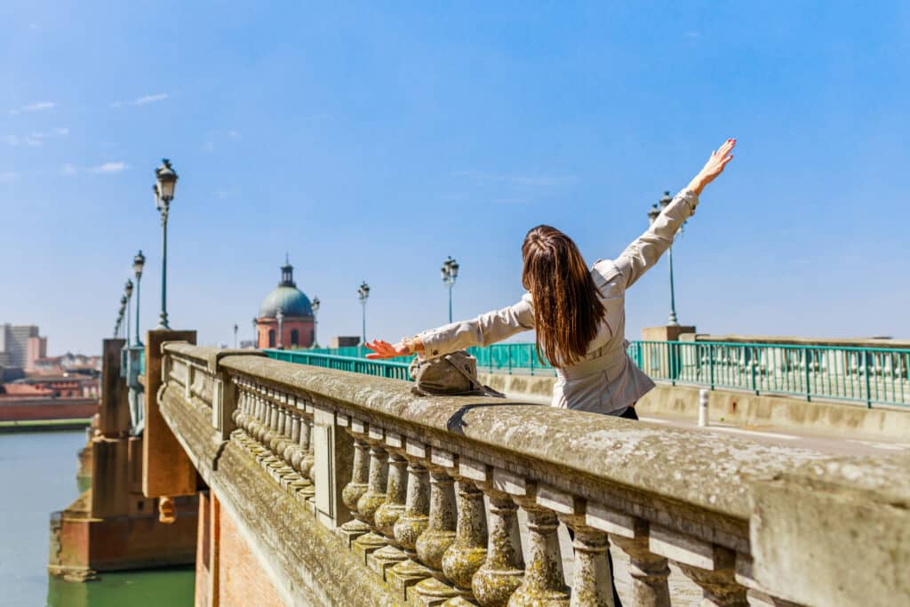 Toulouse: Femme aux bras ouverts sur le pont, vue sur la Garonne Femme les bras ouverts sur un pont en pierre. Vue sur le dôme de la Grave de Toulouse et la Garonne sous ciel bleu.